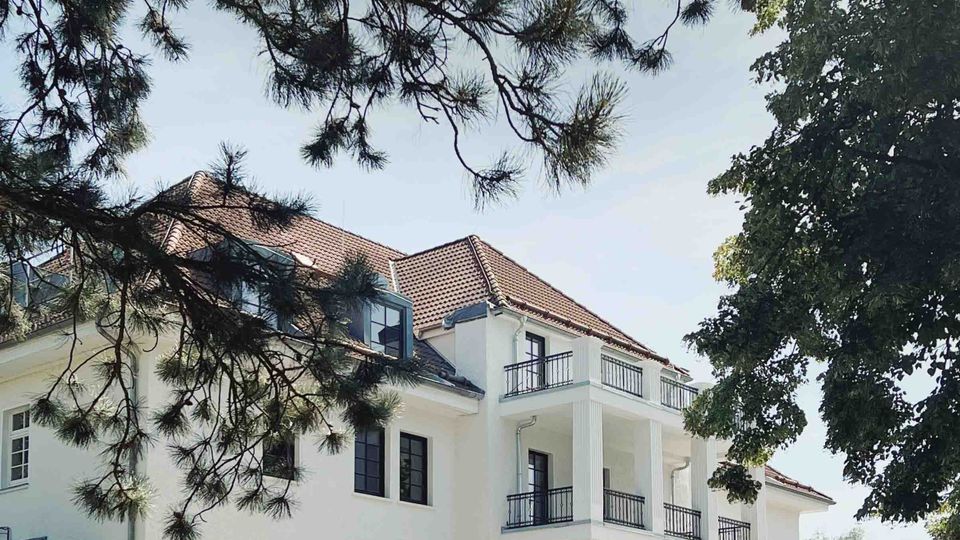 Ein weißes, dreistöckiges Gebäude mit einem roten Ziegeldach und Balkonen an der rechten Seite, umgeben von Bäumen. Im Vordergrund parken ein klassisches Auto und ein modernes Auto auf einer asphaltierten Fläche.