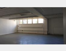 Leerer Raum mit weißen Wänden, einem grauen Boden und einer weißen Rasterdecke mit eingebauten Leuchtstofflampen. Eine Fensterfront mit mehreren Fenstern auf der rechten Wand sorgt für Tageslicht, darunter befinden sich ein Heizkörper und eine Jalousie.