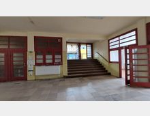 Eingangsbereich eines Gebäudes mit cremefarbenen Wänden, roten Rahmen aus Holz und Glaselementen an Türen und Fenstern. Eine breite Treppe mit dunklem Holz führt zu einer weiteren Glastür, die ins Freie zeigt.