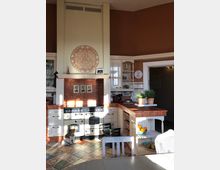 Wohnküche mit rustikalem Stil, ausgestattet mit einem großen weißen Herd und einer Arbeitsplatte aus Terrakotta-Fliesen. Im Raum befinden sich Holzmöbel, dekorative Pflanzen, ein Wandornament und ein Hund, der auf dem gemusterten Fliesenboden sitzt.