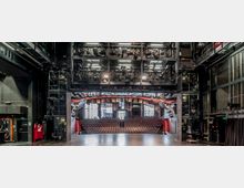 Blick von einer Theaterbühne in den Zuschauerraum eines modernen Theaters mit Reihen bestuhlter Sitzplätze. Die Bühne ist umgeben von schwarzer Bühnentechnik mit zahlreichen Beleuchtungselementen und Stahlkonstruktionen.