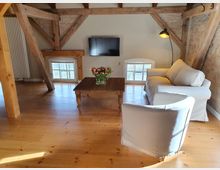 Gemütliches Wohnzimmer in einem Dachgeschoss mit sichtbaren Holzbalken, Backsteinwand und schrägen Wänden. Das Zimmer enthält eine helle Couch, einen Sessel, einen Holztisch mit einem Tulpenstrauß, kleine halbhohe Fenster und einen an der Wand montierten Fernseher.