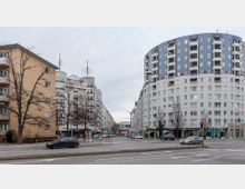 Straßenszene in einer Stadt mit mehrstöckigen Wohngebäuden, darunter links ein beige-gelbes Gebäude und rechts ein modernes, teils blaues Gebäude mit abgerundeter Form. Im Vordergrund eine Straßenkreuzung mit Autos und kahlen Bäumen.