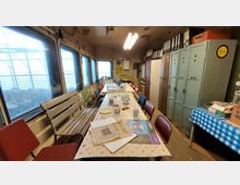 Ein einfacher Aufenthaltsraum in einem industriellen oder historischen Setting mit einer langen Tischreihe, umgeben von roten Stühlen und einer Holzbank. Im Hintergrund befinden sich Metallspinde, Aktenordner und eine Wand mit Fenstern, die teilweise vergittert sind und Tageslicht hereinlassen.