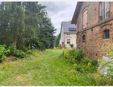 Rasenfläche zwischen einer roten Backsteinfassade rechts und dichten Nadelbäumen links, im Hintergrund ein weißes Haus mit gelben Fenstereinfassungen und grauem Dach; umgeben von Sträuchern und Wildpflanzen. Der Himmel ist bewölkt.