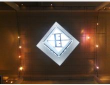 Blick auf eine Decke mit einem großen, rautenförmigen Glaselement, das Tageslicht hereinlässt. Umgeben ist das Element von Wänden und einer Struktur mit mehreren kleinen, warm leuchtenden Lichtern.