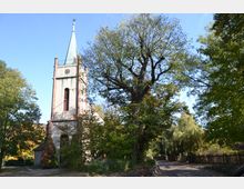 Kirche mit hellem Turm und grünlicher Spitze, umgeben von Bäumen und einem gepflasterten Weg im Vordergrund. Die Umgebung zeigt eine ländliche, bewaldete Szenerie bei sonnigem Wetter.