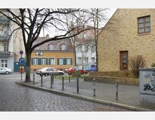 Straßenszene mit gepflastertem Gehweg, einem kleinen Platz mit Autos und mehreren Gebäuden im Hintergrund, darunter ein gelbes Haus mit grünen Fensterläden. Im Vordergrund steht ein Baum, und rechts ist eine Fassade aus gelbem Ziegelstein zu sehen.