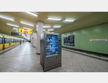 U-Bahnhof mit gelber U-Bahn auf der linken Seite, einem Snackautomaten in der Bildmitte und grünen gefliesten Wänden. Ein Schild mit der Aufschrift „Kurfürstendamm" und verschiedene Hinweisschilder sind an den Wänden und der Decke angebracht.