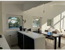 Heller Wohnraum mit minimalistischem Design, bestehend aus einem schwarzen Esstisch mit Stühlen, einer weißen Theke und mehreren großen Fenstern, die Tageslicht hereinlassen und den Blick auf grüne Bäume und benachbarte Gebäude ermöglichen. Im Hintergrund eine helle Sofaecke mit Kissen.