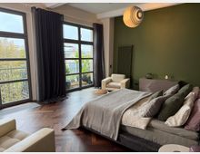 Modernes Schlafzimmer mit großen bodentiefen Fenstern, verdunkelt durch dunkle Vorhänge, einer olivgrünen Wand und Fischgrätparkettboden. Ein Doppelbett mit grauer Bettwäsche, zwei Sesseln und eine Kommode sind sichtbar, daneben ein Balkon mit Pflanzen.