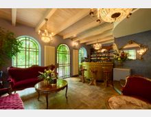 Gemütlicher Innenraum eines Cafés oder einer Lounge mit vintage-inspirierter Einrichtung. Zu sehen sind Sessel und Sofas mit rotem Samtbezug, ein goldener Bartresen mit Hockern, große Fensterbögen mit Blick ins Grüne und dekorative Kronleuchter an einer Decke mit Holzbalken.