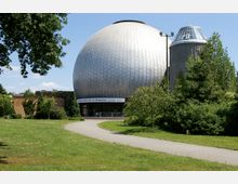 Kuppelförmiges Planetarium mit silberner Metallfassade, flankiert von einem weiteren zylindrischen Gebäude mit gläserner Spitze, umgeben von grünen Wiesen, Bäumen und einem gewundenen Gehweg im Vordergrund.