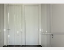 Innenraum mit einer schlichten weißen Tür in der Mitte, flankiert von glatten Glasflächen mit vertikalen Linien und Spiegelreflexionen; dunkler Fußboden und minimalistische Gestaltung.