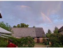 Ein ländlicher Hof mit einer großen Scheune aus Ziegeln und Wellblechdach, umgeben von Bäumen und Büschen. Am Himmel ist ein Regenbogen erkennbar, während eine Person im roten Shirt links im Vordergrund steht.