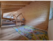Holzgetäfeltes Dachgeschosszimmer mit schrägen Deckenbalken, einem Kinderbett in Hausform, einem größeren Bett und einem bunten Spielteppich mit Straßenmotiven auf dem Holzboden. Tageslicht scheint durch ein schräges Fenster am linken Rand.