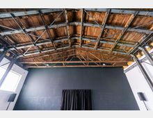 Ein Innenraum mit einer sichtbaren Holzdecke, die durch eine Metallkonstruktion gestützt wird. Die Wände sind in Weiß und Dunkelgrau gehalten, und an der grauen Wand hängt ein schwarzer Vorhang; rechts und links sind Lautsprecher erkennbar.