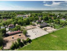 Luftaufnahme eines Reiterhofs in einer ländlichen Umgebung. Zu sehen sind Reitplätze, Stallgebäude, Wohnhäuser und umgebendes Grünland mit Bäumen und Feldern im Hintergrund.