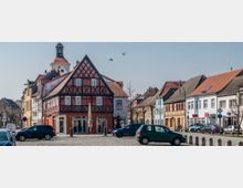Historisches Fachwerkhaus mit rotem und schwarzem Fachwerk in einer Kleinstadt, umgeben von pastellfarbenen Gebäuden mit Ziegeldächern; im Hintergrund ist ein Kirchturm mit Uhr zu sehen. Der gepflasterte Platz im Vordergrund ist mit geparkten Autos gesäumt.