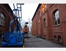 Eine schmale Gasse zwischen zwei roten Backsteingebäuden mit großen Fenstern. Im Vordergrund befindet sich eine blaue Hebebühne, während im Hintergrund eine weitere Hebebühne mit Scheinwerfer sichtbar ist; Kabel und Verkehrshütchen liegen verstreut auf dem Boden.