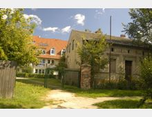 Ein älteres, verwittertes Gebäude mit bröckelnder Fassade steht neben einem moderneren Wohnhaus mit rotem Ziegeldach und weißen Fenstern. Im Vordergrund befindet sich ein grünes Tor, das von einem Ziegelpfeiler und einem Maschendrahtzaun flankiert wird, umgeben von Bäumen und Rasen.