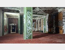 Innenraum eines verlassenen Gebäudes mit abblätternder Farbe an Decke und Wänden, graffitibeschmierten grünen Stützpfeilern und rotem Teppichboden. An der rechten Seite stehen Fensterreihen und Heizkörper, im Hintergrund führen offene Durchgänge in weitere dunkle Räume.