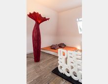 Schlafzimmer mit minimalistischem Design: ein Futonbett mit orangefarbener Bettdecke und bunten Kissen auf einem Holzboden, eine skulpturale rote Stehlampe links und eine dekorative weiße Schriftplastik mit dem Text „I'D BE SUPER GOOD“ auf einem Sockel im Vordergrund. Im Hintergrund ein kleines, geöffnetes Fenster.