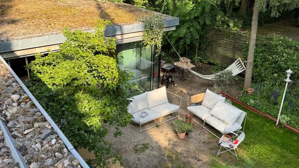 Backyard patio beside a modern, flat-roofed house with large glass doors, outdoor sofas and chairs on a sandy terrace, and a hammock near a wooden fence. The garden is bordered by tall, dense trees and shrubs, with a small lawn area in the foreground.