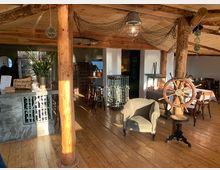 Gemütlicher Innenraum eines Cafés oder Restaurants mit maritimem Dekor, darunter Holzbalken, ein Schiffssteuerrad, Fischernetze an der Decke und Holzboden. Ein Tresen aus Stein und Holzmöbel ergänzen das rustikale Ambiente.