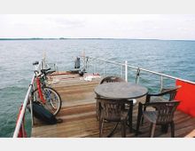 Holzterrasse eines Bootes mit Blick auf eine weite Wasserfläche; im Vordergrund steht ein runder Plastiktisch mit Stühlen, daneben sind zwei Fahrräder an der Reling befestigt. Im Hintergrund sind Seile und andere Bootszubehörteile sichtbar.