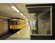 Gelber U-Bahn-Zug in einer unterirdischen U-Bahn-Station, links einfahrend, mit leeren Sitzplätzen entlang des rechten Bahnsteigs; im Vordergrund eine verglaste Kabine mit Knöpfen und einem Mikrofon.