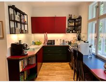 Moderne Küche mit roten und grünen Fronten, offenen Regalen und Holzfußboden. Eine Arbeitsplatte mit Küchengeräten, ein Gasherd, eine Spüle und ein Esstisch vor einem großen Fenster prägen den Raum.