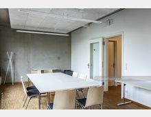 Moderner Besprechungsraum mit weißen Wänden und einer grauen Betonwand, ausgestattet mit einem weißen rechteckigen Tisch und mehreren Holzstühlen mit schwarzen Sitzflächen. Der Raum hat einen Holzboden, rechteckige LED-Deckenlampen und eine Glastür, die in einen angrenzenden Flur führt.