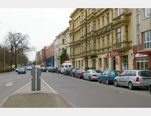 Straßenszene mit mehrstöckigen, historischen Wohngebäuden auf der rechten Seite, darunter ein Geschäft mit Apothekenzeichen. Links führt eine mehrspurige Straße mit parkenden Autos, kahlen Bäumen und einem schmalen Mittelstreifen entlang.