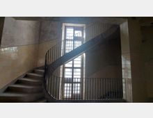 Spiralformige Treppe in einem Gebäude mit schmiedeeisernem Geländer, flankiert von beige gekachelten Wänden; im Hintergrund ein großes Fenster mit Holzrahmen, das Tageslicht hereinlässt.