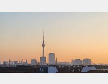 Stadtansicht von Berlin bei Sonnenuntergang mit dem Fernsehturm im Zentrum, umgeben von modernen Gebäuden und Wohnhäusern im Vordergrund. Der Himmel ist in warmen Orange- und Blautönen gefärbt.