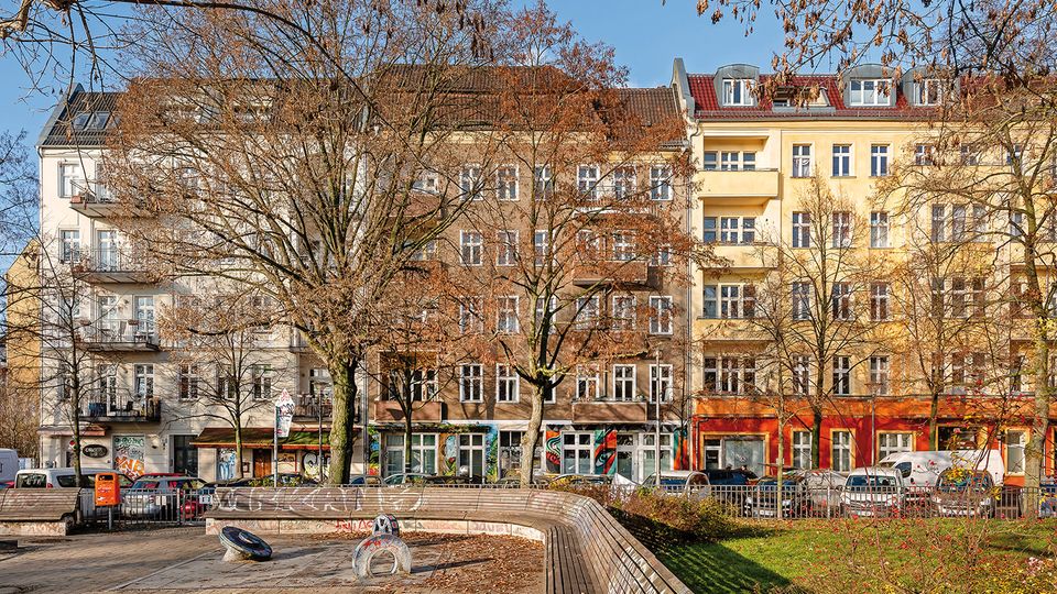 Blick auf einen kleinen Stadtplatz mit gebogener Holzsitzbank, gepflasterter Fläche und Rasenbeet mit kahlen Bäumen. Im Hintergrund stehen mehrstöckige Altbau-Wohnhäuser mit vielen Fenstern, Balkonen und farbigen Erdgeschosszonen entlang einer Straße mit geparkten Autos.