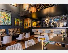 Elegante Bar mit modernem Design; vorne befinden sich Tische mit grauen Polsterstühlen und floralen Sitzbänken, im Hintergrund eine Theke mit goldfarbenen Barhockern und dekorativen Lampen, sowie bunte abstrakte Wandgemälde.