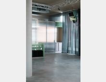 Minimalistischer Innenraum mit grauem Betonboden, einer grünen, gefliesten Theke und Metalltüren im Hintergrund. Ein Vorhang bedeckt teilweise die Fenster, und an der Decke sind Traversen sowie Lautsprecher angebracht.