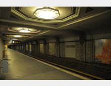 U-Bahn-Station mit rechteckigem Bahnsteig, gekachelten Wänden und dekorativen Leuchten an der Decke. Auf den Wänden befinden sich bunte Wandbilder, und ein Schild mit der Aufschrift „Breitenbach“ ist sichtbar.