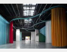 Ein großer, moderner Innenraum mit glänzendem Betonboden, farbigen Vorhängen in Blau, Rot, Weiß und Orange, die von einer wellenförmigen Metallstruktur an der dunklen Decke hängen. Im Hintergrund ist eine Türöffnung mit Tageslichteinfall sichtbar.