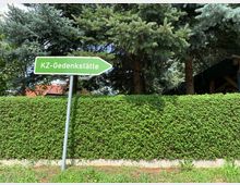 Grünes Straßenschild mit der Aufschrift „KZ-Gedenkstätte“ und einem Pfeil nach rechts, vor einer Hecke und hohen Nadelbäumen. Im Hintergrund ist teilweise ein Ziegeldach sichtbar.