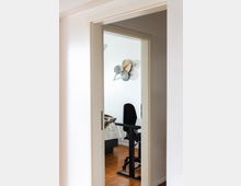 Blick durch eine offene Tür in ein Zimmer mit einem höhenverstellbaren schwarzen Schreibtisch und einem schwarzen Bürostuhl, Holzboden und weißer Wand mit dekorativen Wandornamenten in Blattform. Im Hintergrund ist ein Bett mit weißer Tagesdecke und Fellimitat-Decke sichtbar.