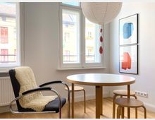 Heller Essbereich in einer Wohnung mit rundem Holztisch, kleinen Hockern und einem Sessel, daneben große hohe Fenster mit Blick auf gegenüberliegende Wohnhäuser. An der Wand hängen zwei gerahmte abstrakte Drucke, und von der Decke hängt eine große Papierlampion-Leuchte über dem Tisch.