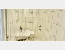 Weiß gefliestes Badezimmer mit einem Waschbecken, einem kleineren Wandspiegel, einer Toilette mit eingebautem Spülkasten und einem Toilettenpapierhalter an der rechten Wand. Der Boden ist mit weißen Fliesen im diagonalen Muster verlegt.