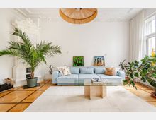 Helles Wohnzimmer mit weißer Stuckdecke, Parkettboden und großem Fenster mit Vorhängen. Vor einer hellblauen Couch stehen ein Glas-Couchtisch und ein heller Teppich; links ist ein weißer Kachelofen, dazu mehrere große Zimmerpflanzen und zwei gerahmte Bilder an der Wand.