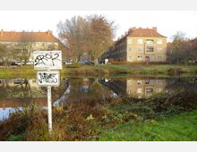 Mehrfamilienhäuser mit beige-brauner Fassade und roten Ziegeldächern stehen an einem kleinen Teich, in dem die Gebäude und Bäume klar reflektiert werden. Im Vordergrund befinden sich ein mit Graffiti beschmiertes Verbotsschild und eine Wiese mit niedrigem Bewuchs.