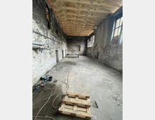 Großer, leerer Innenraum eines alten Industriegebäudes mit unverputzten Ziegelwänden, hohen Fenstern und neuem Holzbalkendach. Der Betonboden ist teilweise mit Schläuchen, Kabeln und einer Holzpalette bedeckt.