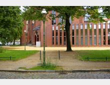 Backsteinbau mit neugotischen Elementen, darunter ein spitzbogiges Portal, flankiert von modernen schmalen Glas- und Backsteinsäulen. Im Vordergrund befinden sich ein Gehweg, Rasenflächen, Bäume und eine historische Straßenlaterne.