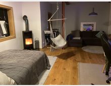 Wohnraum mit Holzboden und moderner Einrichtung. Im Vordergrund befinden sich ein graues Bett, ein hängender Hängesessel und ein Holzofen, im Hintergrund ein dunkles Ecksofa, ein Wandbild und eine Hängeleuchte.